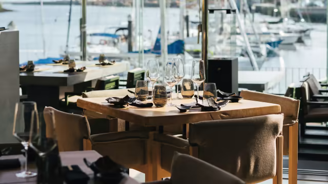 Interior de restaurante moderno com área de refeições, mesas com copos dispostos, cadeiras estofadas confortáveis e vista pitoresca de barcos e água.