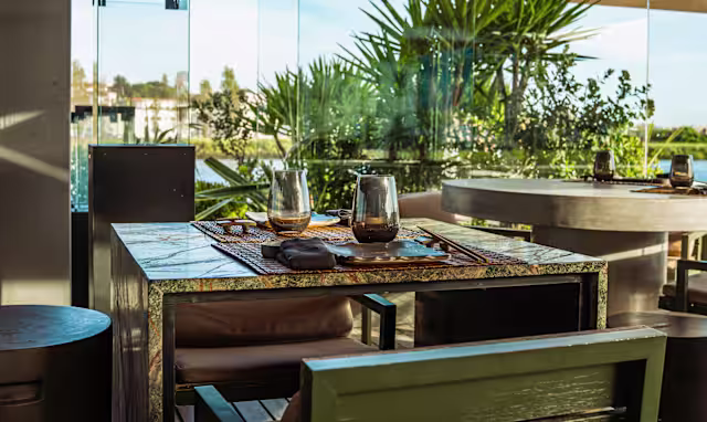 Interior de um restaurante moderno com uma mesa elegante para dois, rodeada de luz natural e vegetação visível através de grandes janelas de vidro, criando uma atmosfera relaxante e sofisticada.