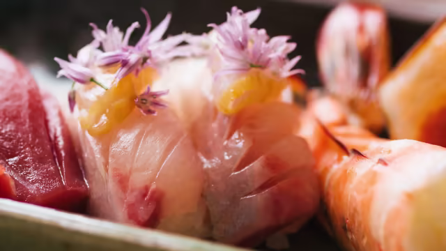 Uma seleção de sushi com fatias de peixe fresco, incluindo sashimi delicado de atum e outro peixe branco, decorado com flores comestíveis. Acompanha camarões suculentos, com cascas brilhantes, destacando a frescura do prato.