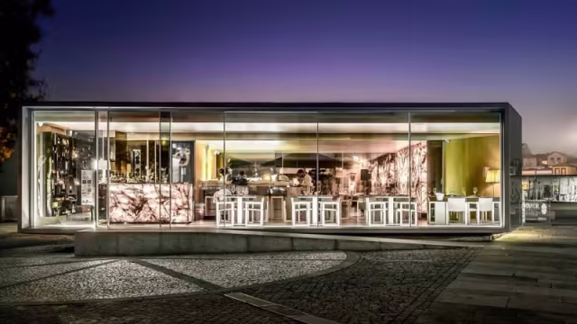 Restaurante moderno com exterior em vidro, interior contemporâneo e mobiliário minimalista, iluminado de forma acolhedora, criando uma atmosfera convidativa.