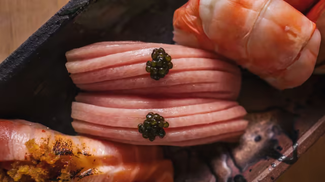 Fatias de sushi elegantemente dispostas, com peixe rosa delicado, possivelmente atum ou salmão, cobertas com pequenas porções de caviar e acompanhadas por camarões frescos e brilhantes.