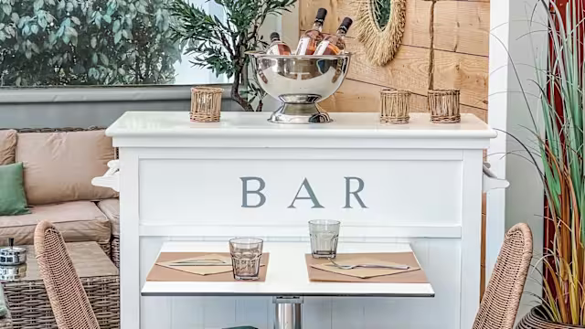 Espace bar chaleureux avec un comptoir blanc, des bouteilles de vin dans un seau à glace, et une ambiance accueillante agrémentée de verdure.