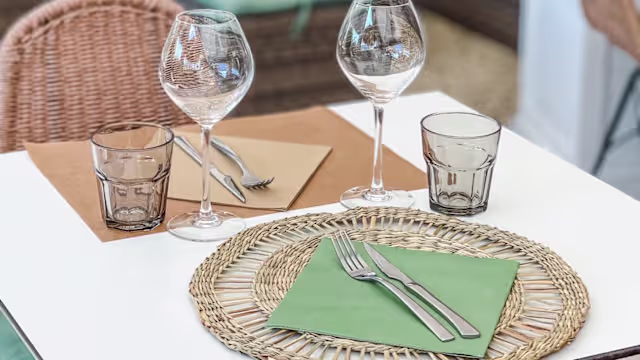 Table élégamment dressée avec des verres à vin et un tumbler, nappes vertes pliées, et couverts prêts à l'emploi, évoquant une expérience culinaire décontractée mais raffinée.