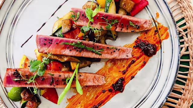 Trois tranches de poisson poêlé, probablement du thon, garnies de jeunes pousses et nappées d'une sauce sombre, accompagnées d'un mélange coloré de légumes sautés et d'une purée d'orange lisse.