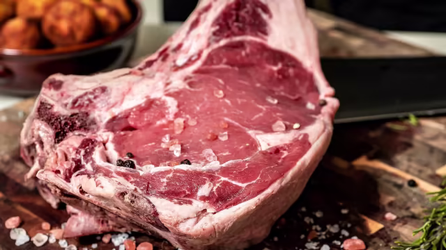 Taglio di carne crudo, probabilmente ribeye o tomahawk, con marmorizzazione ricca e condimenti di sale grosso e pepe.