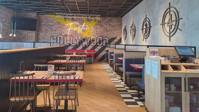 Interior moderno con elementos industriales y casuales, paredes de ladrillo expuesto y gráficos audaces, asientos en booths rojos y sillas de madera, iluminación cálida y elementos decorativos contemporáneos.