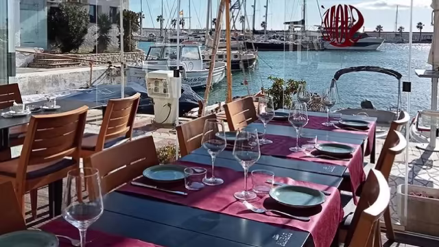 Área de comedor al aire libre junto al agua, con mesas cubiertas con manteles rojos y copas de vino elegantes, creando un ambiente relajado y agradable.