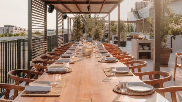 Área de comedor al aire libre con una larga mesa de madera, rodeada de sillas, decorada con platos y copas, creando un ambiente acogedor y elegante.