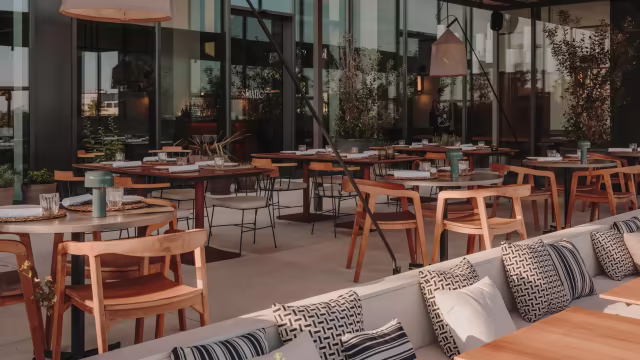 Interior de restaurante moderno con área exterior espaciosa, mesas de madera y sillas de metal negro, creando un ambiente contemporáneo y acogedor.