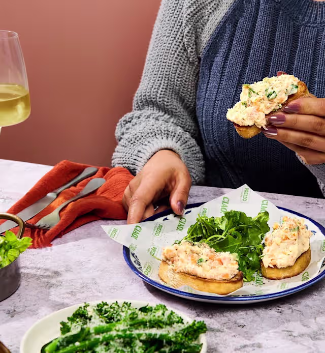 Toasted bread topped with a creamy seafood spread, garnished with fresh herbs, served with a side of leafy greens.