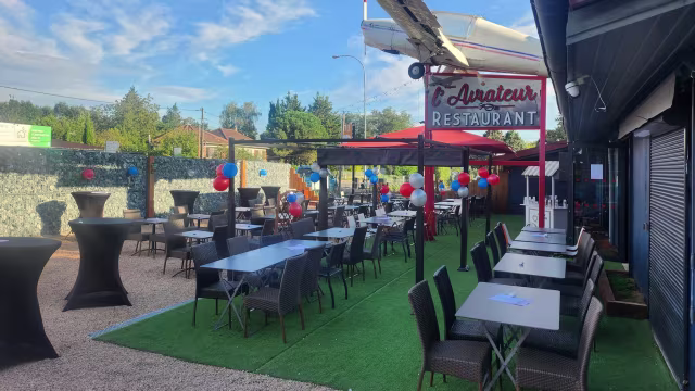 Espace de restauration en plein air avec tables sur une surface herbeuse, chaises sombres et nappes blanches, décoré de ballons colorés et bien éclairé par la lumière naturelle.