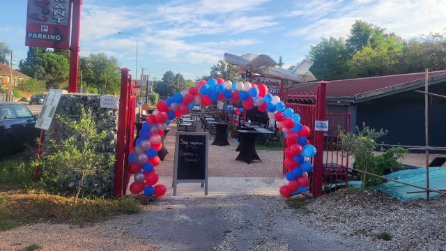 Entrée d'un restaurant décorée d'un arc festif en ballons rouges, blancs et bleus, avec un chemin en gravier menant à un panneau d'affichage, et des tables installées pour un espace de restauration en plein air.