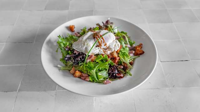 Salade fraîche composée de mélanges de feuilles vertes, surmontée d'un œuf poché, de morceaux de bacon croustillants et de croûtons, le tout agrémenté d'une vinaigrette et de ciboulette finement hachée.
