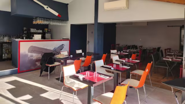 Intérieur moderne et décontracté avec des chaises colorées, des sets de table rouges et un bar orné d'une fresque d'avion, créant une atmosphère accueillante et spacieuse.