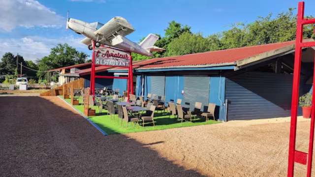 Restaurant au charme rustique avec une façade bleue et un toit rouge, ornée d'un avion vintage au-dessus de l'entrée, créant une atmosphère décontractée et accueillante.