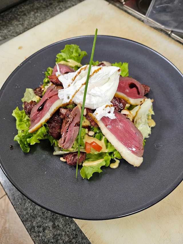 Salade fraîche avec des tranches de poitrine de canard tendre sur un lit de laitue croquante, garnie d'un œuf poché, de tomates hachées et de pignons de pin grillés, le tout rehaussé d'une vinaigrette.