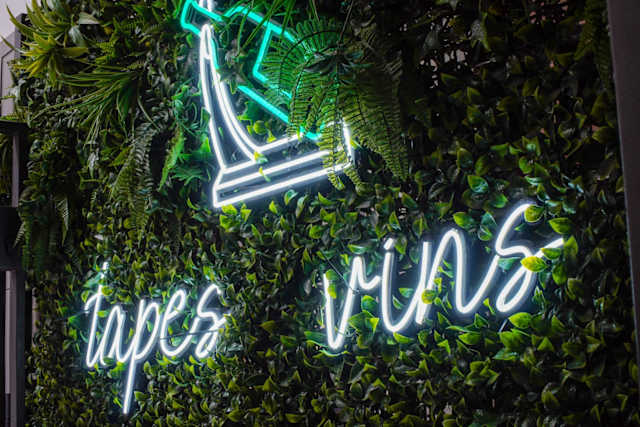 Cartel de neón que dice "tapes" y "vins" iluminado en una pared verde cubierta de plantas, creando un ambiente moderno y acogedor de bar de vinos.