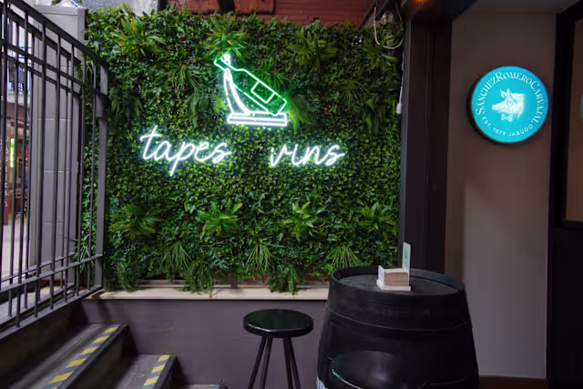 Entrada de restaurante encantadora con una pared verde cubierta de plantas, un letrero de neón "tapes vins" y un barril de madera decorativo.