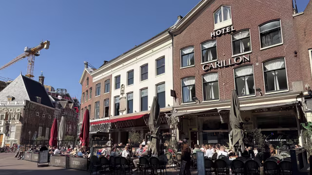 Een levendige restaurantgevel van Hotel Carillon met drukke buitenzitplaatsen waar gasten in de zon van hun maaltijden genieten. Het gebouw heeft een klassieke architectuur met grote ramen en een opvallend bord. Parasols bieden schaduw voor de tafels, terwijl een bouwkraan op de achtergrond wijst op de ontwikkeling in de omgeving. De algehele sfeer straalt levendigheid en sociale interactie uit.