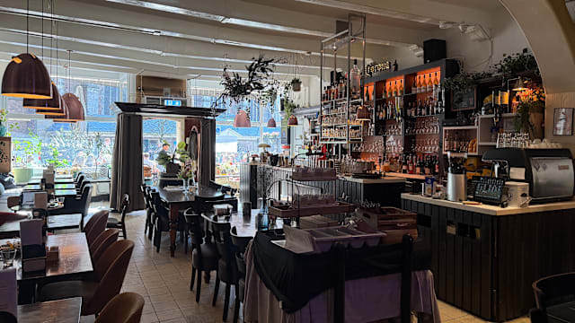 Een helder en uitnodigend restaurantinterieur met een mix van houten tafels en stoelen, verlicht door warme hanglampen. Een goed gevulde bar is zichtbaar op de achtergrond, omringd door groen en decoratieve elementen. Grote ramen laten natuurlijk licht binnen, wat de gastvrije sfeer versterkt.