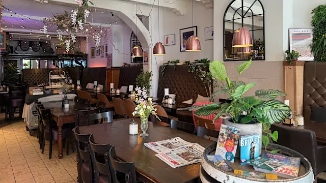 Het interieur van het restaurant is gezellig en uitnodigend, met houten tafels en stoelen voor een ontspannen eetervaring. Zachte verlichting van hanglampen zorgt voor een warme gloed, terwijl decoratieve elementen zoals potplanten en bloemstukken de natuurlijke sfeer versterken. De wanden zijn versierd met ingelijste kunstwerken, wat bijdraagt aan de charme van het restaurant. De indeling suggereert een verwelkomende omgeving voor zowel casual diners als kleine bijeenkomsten.