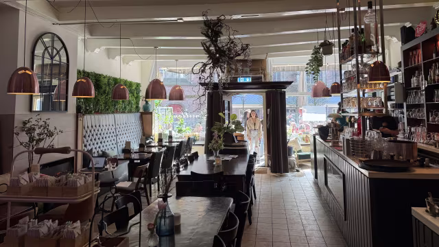 Ruime restaurantinterieur met een mix van houten en metalen meubels, hangende koperen lampen en weelderige planten aan de muren, wat een frisse en uitnodigende sfeer creëert. Grote ramen zorgen voor veel natuurlijk licht, terwijl een stijlvolle bar met glazen in de achtergrond zichtbaar is. Het ontwerp combineert moderne elementen met een gezellige, verwelkomende uitstraling.