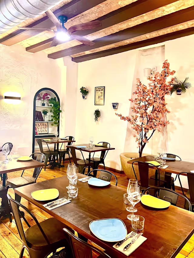 Interior de restaurante acogedor con vigas de madera, iluminación cálida, mesas de madera y sillas de metal negro, decorado con plantas y arte en las paredes, destacando un árbol de cerezo.