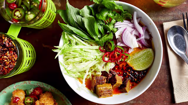 A bowl of pho with aromatic broth, tender beef, fresh herbs, crisp lettuce, red onions, and green chili slices, served with a lime wedge.