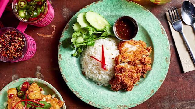 Crispy fried chicken coated in golden breadcrumbs, served with fluffy white rice, fresh cucumber slices, leafy greens, and a sweet and spicy dipping sauce, garnished with red chili.