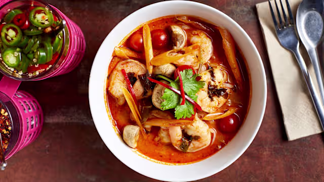 Aromatic shrimp soup with plump shrimp, sliced mushrooms, and cherry tomatoes in a rich, reddish-orange broth, garnished with fresh cilantro and red chili.