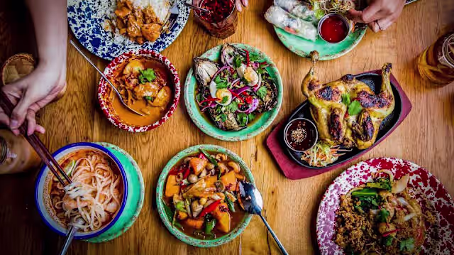 A whole chargrilled chicken served with a fresh herb salad, accompanied by creamy coconut curry with vegetables, a herb-garnished noodle dish, and a tropical fruit salad, creating a vibrant communal dining experience.