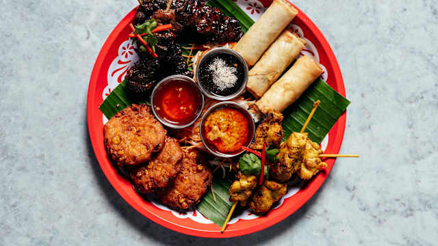 A vibrant platter of appetizers featuring golden-brown spring rolls, crispy fritters, and skewers of chargrilled meat, accompanied by dipping sauces.