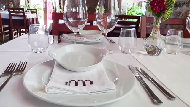 Mesa de jantar bem arrumada, com um prato branco ao centro, um guardanapo dobrado com um logótipo e uma pequena taça, sugerindo um molho ou aperitivo. A mesa inclui copos de vinho e água polidos, e uma simples arranjo floral que acrescenta cor e frescura ao ambiente.
