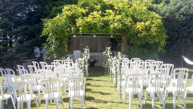 Installation de mariage en plein air avec une arche décorée de tissu doux, entourée de verdure et de fleurs, et des chaises blanches disposées sur une pelouse.