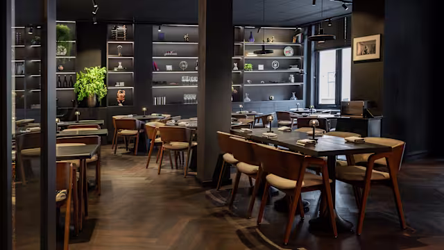 Een modern restaurantinterieur met donkere muren en houten vloeren, verlicht door zachte verlichting voor een intieme sfeer. Strakke houten tafels zijn ingericht met minimalistische decoratie en stijlvolle stoelen passen bij het eigentijdse ontwerp. Planken op de achtergrond tonen verschillende decoratieve items, wat een vleugje verfijning toevoegt, terwijl potplanten een hint van groen bieden.