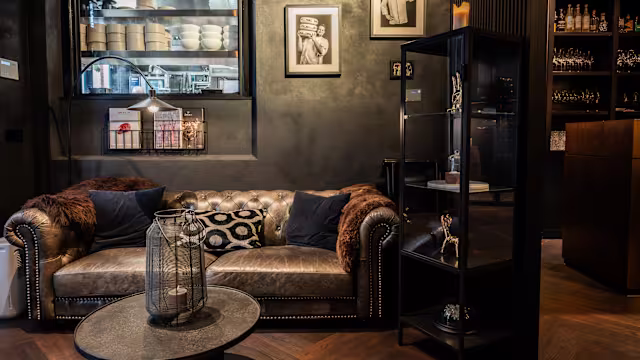 Stijlvol restaurantinterieur met een luxe, donkerleren bank en decoratieve kussens. De muren zijn in een diepe kleur geschilderd, versierd met ingelijste zwart-witfoto's. Een moderne boekenkast toont diverse items, terwijl een ronde salontafel de gezellige en verfijnde sfeer versterkt. Zachte verlichting van een lamp creëert een warme ambiance.