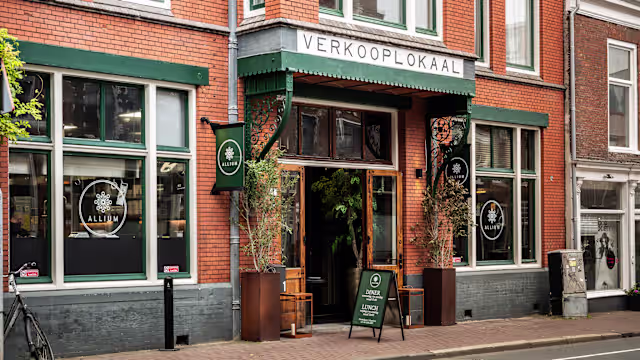 Charmante restaurantgevel met een bakstenen façade en grote ramen die uitzicht op het interieur bieden. De ingang is gemarkeerd met een bord 'VERKOOPLOKAAL' en twee zwarte banners met een logo. Potplanten flankeren de ingang en een klein krijtbord staat op de stoep, waarschijnlijk met het menu of specials. De algehele uitstraling is uitnodigend en goed onderhouden, wat een gastvrije sfeer voor dinergasten suggereert.