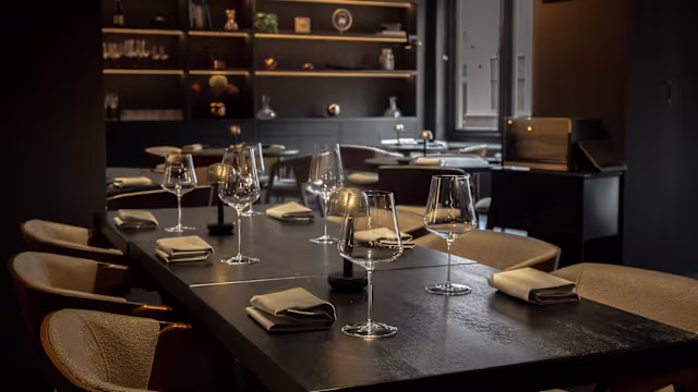 Een modern restaurantinterieur met donkere houten tafels en elegante, minimalistische decoratie. De ruimte is sfeervol verlicht, wat een warme en uitnodigende sfeer creëert. Elk tafeltje is netjes gedekt met glaswerk en gevouwen servetten, terwijl planken op de achtergrond decoratieve items tonen. Het ontwerp benadrukt verfijning en comfort, wat het tot een aantrekkelijke eetomgeving maakt.