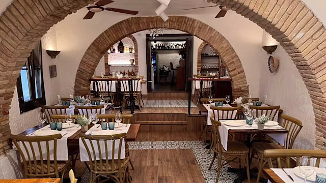 Interior de restaurante acogedor con tonos cálidos de madera, paredes de ladrillo arqueadas y mesas de madera con manteles blancos y cristalería azul. Suelo con diseño de azulejos y buena iluminación.