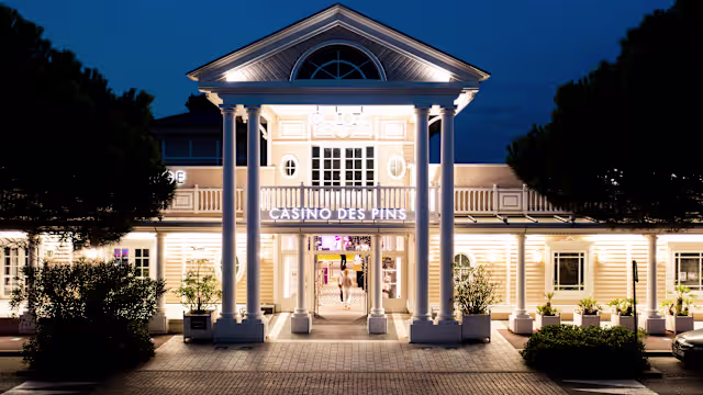 Extérieur du restaurant "Casino des Pins" avec une architecture classique, une entrée encadrée de colonnes blanches, des couleurs douces, de grandes fenêtres et des plantes bien arrangées, créant une ambiance chaleureuse.