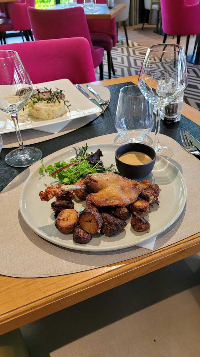 Plat joliment présenté avec une cuisse de poulet croustillante, accompagnée de pommes de terre rôties caramélisées et garnie de légumes frais, le tout servi avec un bol de sauce crémeuse.