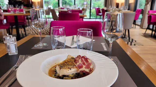 Poisson parfaitement cuit, recouvert d'une croûte dorée de chapelure et d'herbes, servi sur un lit de légumes sautés, accompagné d'une tranche de viande séchée et rehaussé d'une sauce.