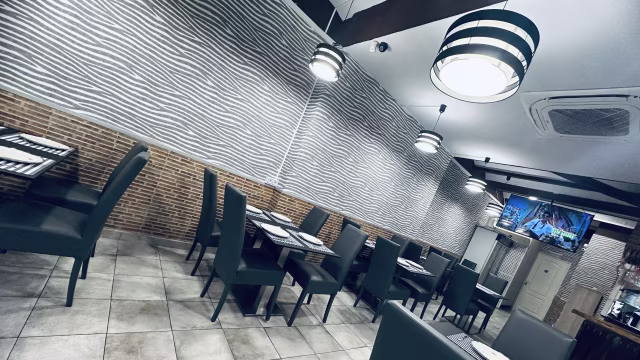 Interior moderno con paneles de pared texturizados en tonos claros, sillas tapizadas en verde oscuro y mesas sencillas. Iluminación con lámparas colgantes a rayas en blanco y negro, creando un ambiente acogedor y casual.