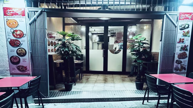 Entrada de un restaurante moderno y acogedor, con puertas de vidrio, plantas en macetas y mesas rojas con sillas negras, ideal para una comida informal.