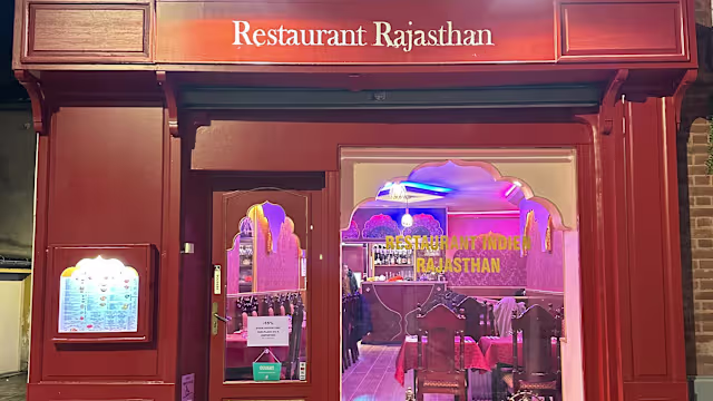 L'extérieur du "Restaurant Rajasthan" présente une façade rouge chaleureuse avec une arche ornée, évoquant des influences architecturales traditionnelles. L'enseigne illuminée et l'éclairage coloré créent une atmosphère vivante, tandis qu'un tableau de menu à gauche met en avant une cuisine indienne authentique.