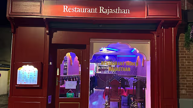 L'extérieur du "Restaurant Rajasthan" présente une façade chaleureuse en tons rouges, avec un nom élégamment affiché au-dessus de l'entrée. De grandes fenêtres laissent entrevoir un intérieur cosy au décor traditionnel, encadré par des arches décoratives, évoquant un thème culturel authentique. Un tableau de menu à gauche propose une sélection de plats inspirés de la cuisine indienne, suggérant une atmosphère accueillante pour les convives.