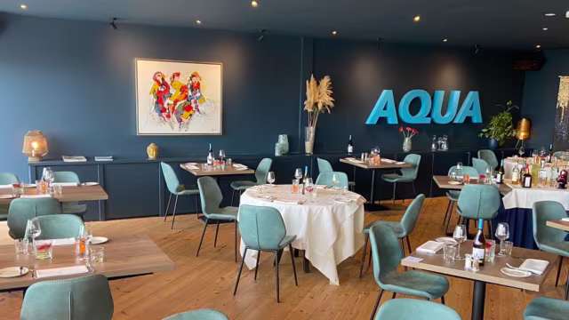 Het restaurant heeft een modern en uitnodigend interieur met een rustgevende blauwe muur en een grote "AQUA" sign. De ruimte is goed verlicht en biedt een warme sfeer, met tafels voorzien van witte tafelkleden en netjes gerangschikt bestek. De stoelen zijn bekleed met zacht, gedempt groen stof, en decoratieve elementen zoals stijlvolle lampen en potplanten voegen elegantie toe.