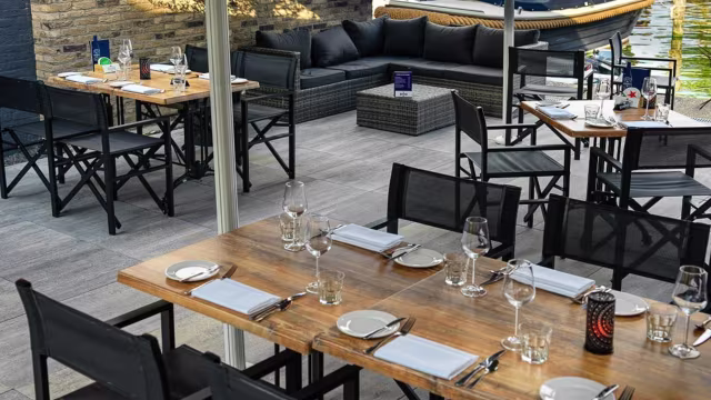 Buiten eetgedeelte met een moderne en uitnodigende sfeer, voorzien van houten tafels, zwarte stoelen en een gezellige lounge met donkere kussens en een lage tafel, omringd door groen en water.