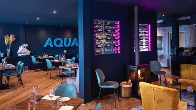 Het restaurantinterieur heeft een moderne en stijlvolle uitstraling met diepblauwe muren en houten vloeren. De mix van teal en mosterdkleurige stoelen creëert een hedendaagse en uitnodigende sfeer. Een prominente wand toont een georganiseerde selectie wijnflessen, verlicht met zachte verlichting. De indeling suggereert een gezellige eetervaring, met tafels voor gasten en een kleine open haard die comfort toevoegt aan de ambiance.
