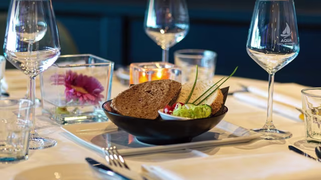 Een kom met donker, rustiek brood in dikke plakken, geserveerd met een romige groene spread van avocado of kruiden, en gegarneerd met blokjes tomaat en fijngehakte ui voor kleur en zuurheid.